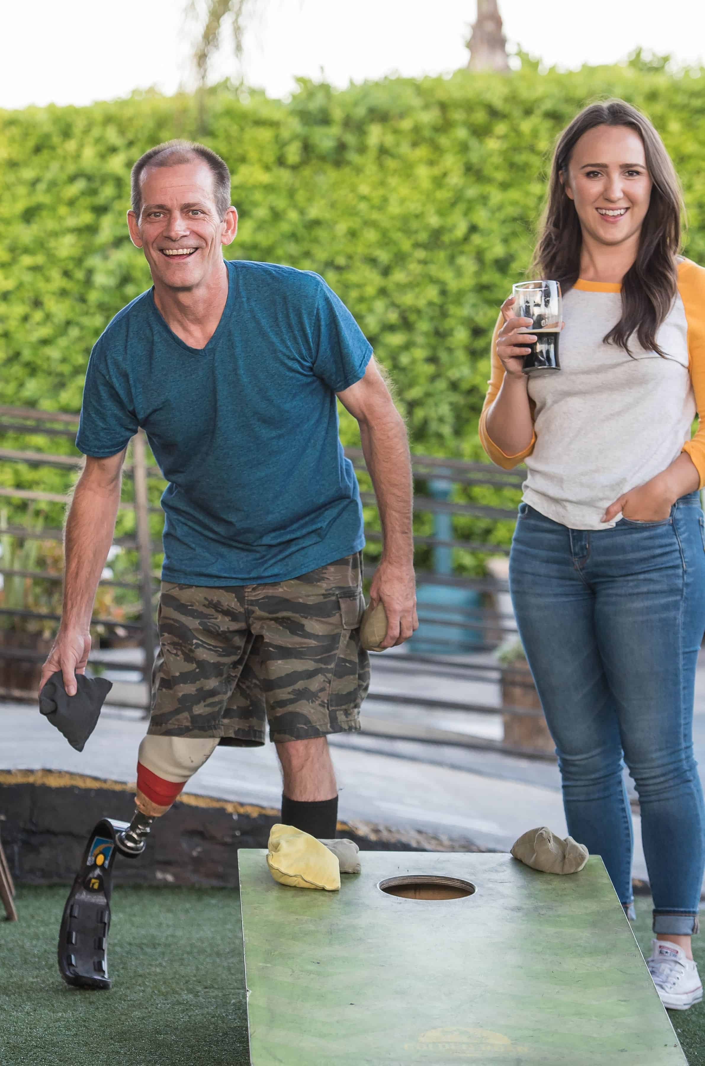 man is playing the best cornhole boards while the girl beside her is drinking a glass of wine while watching him